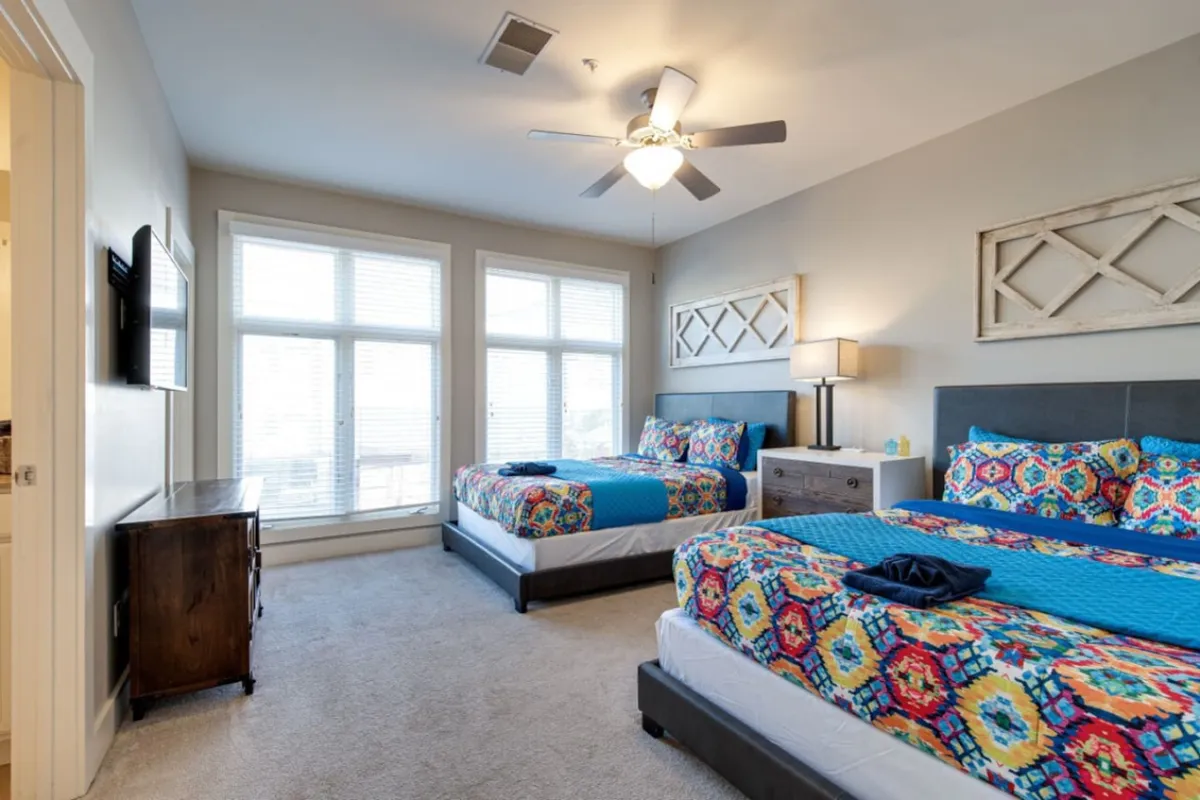 A vibrant bedroom featuring colorful bedding, large windows streaming natural light, a modern lamp, and a cozy nightstand. The room exudes a comfortable, welcoming atmosphere, ideal for relaxing between campus visits or starting the day with morning coffee.