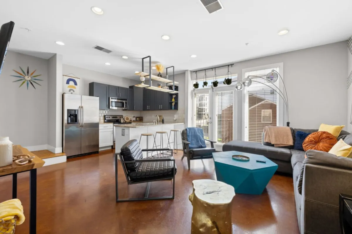 An open-concept area where a comfortable couch, colorful coffee table, kitchen appliances, and decorative wall art are all in view. The living space appears seamlessly connected to the kitchen, emphasizing the flow between lounging and meal prep areas.
