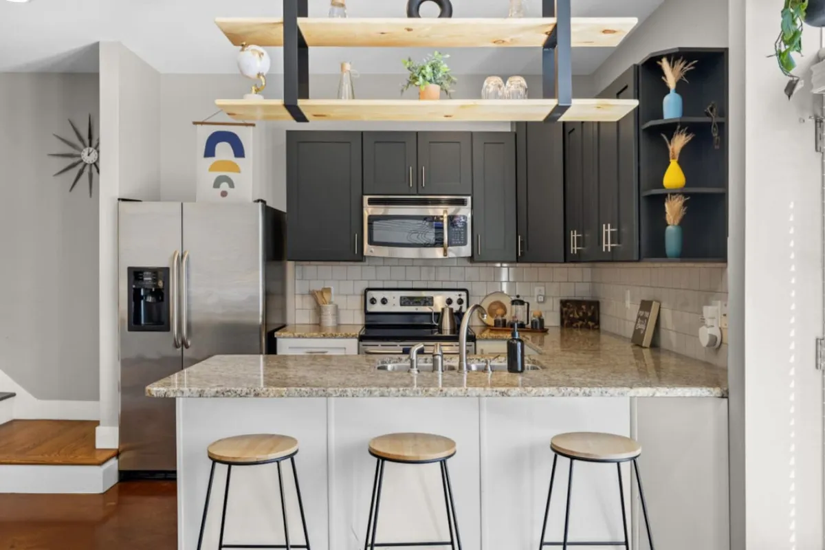 A modern, welcoming kitchen featuring a large island countertop, hanging wooden shelf, black cabinetry, and high-end stainless steel appliances. The space looks ideal for preparing meals, socializing, and enjoying casual dining or coffee breaks.