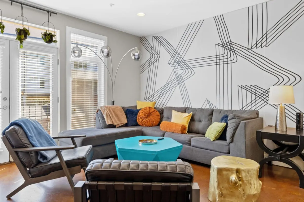 A contemporary living space with colorful pillows on a comfortable sofa, a unique coffee table, geometric wall design, and a stylish floor lamp. The playful décor and open layout suggest a space tailored for both entertaining and relaxing, with ample light giving it a vibrant feel.