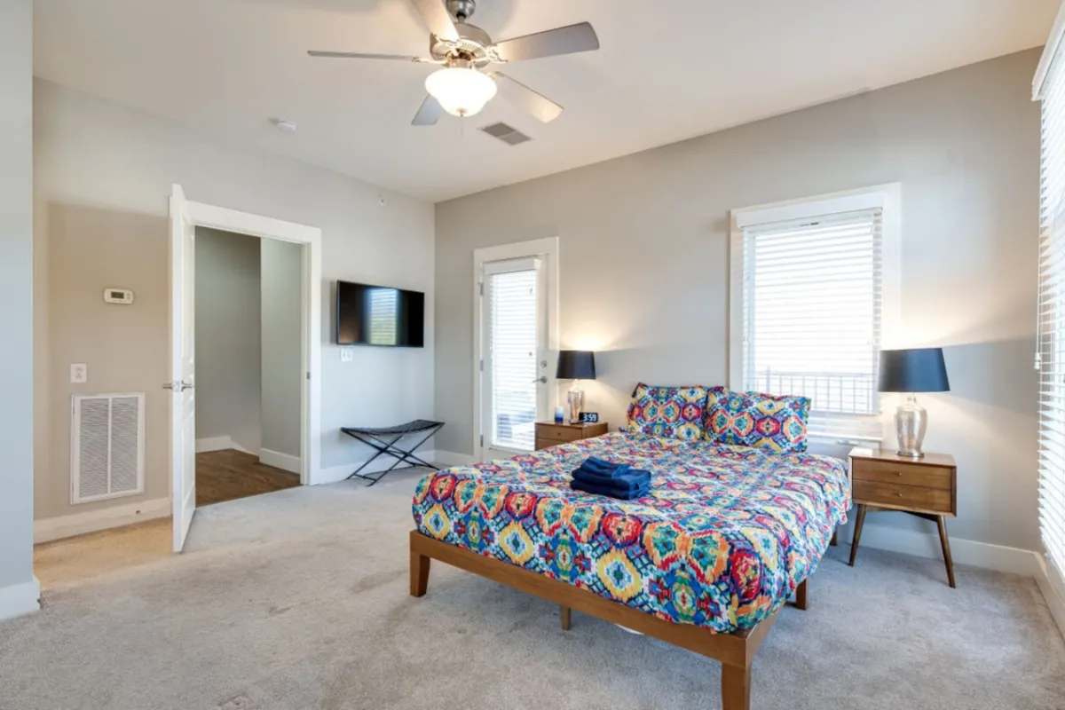 A cozy bedroom scene with a colorful bedspread, a nightstand topped with a lamp, and large windows letting in abundant natural light. The room feels bright and welcoming, suggesting a peaceful morning atmosphere.