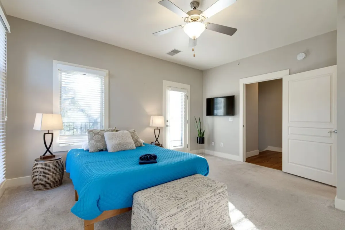 A tranquil bedroom featuring a bed with a bright blue cover, two stylish bedside lamps, a large window with white blinds allowing natural light to fill the space, a modern wall-mounted TV, and a potted plant near the door. The room feels contemporary and restful, inviting guests to relax, watch TV, or start their day refreshed.
