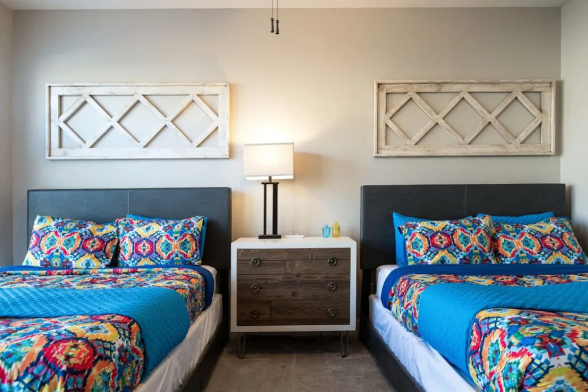 A cozy bedroom scene with colorful bedding, a nightstand and lamp, and decorative wall art. The mood is cheerful, suggesting a welcoming and restful space ideal for both relaxation and socializing. Natural light fills the room, making it feel inviting for a good night's sleep or a leisurely morning.
