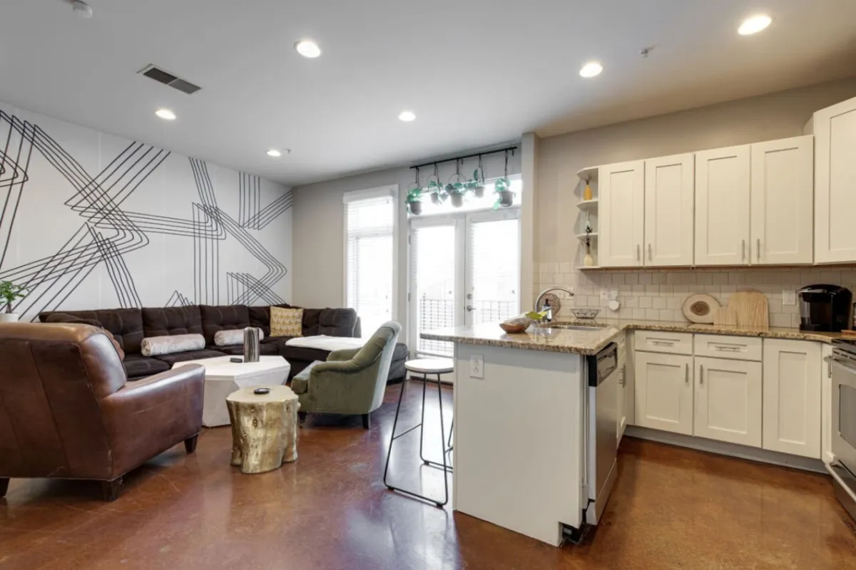 An open-plan space where a kitchen island seamlessly flows into a contemporary dining and living area. The kitchen island, visible alongside a large sectional sofa and artistic wall design, suggests plenty of room for entertaining and shared meals.