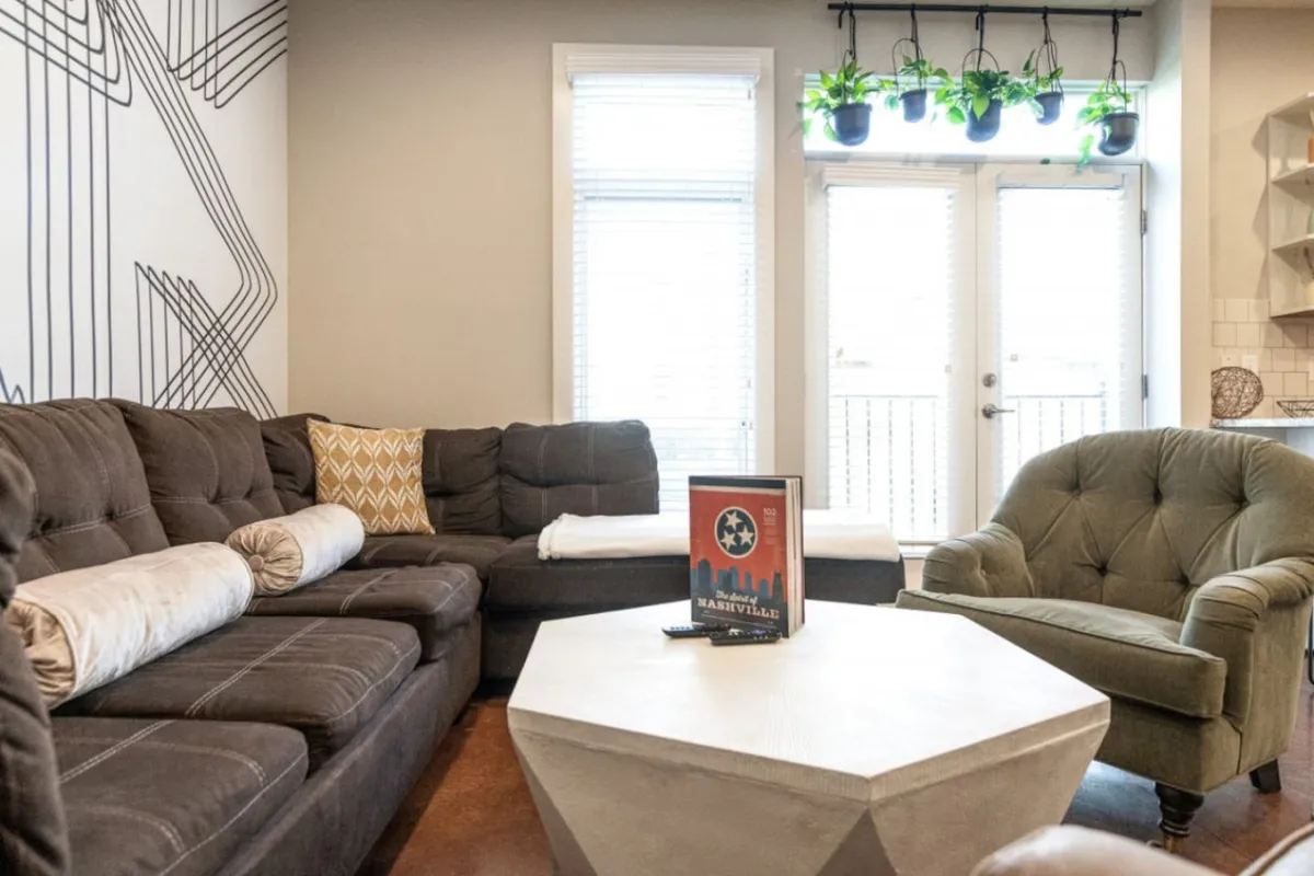 A spacious living room featuring a large sectional sofa positioned around a geometric accent wall and a coffee table with a book. The room is flooded with natural light from windows out of frame, and the overall mood is casual and welcoming—inviting guests to relax, converse, or enjoy a cozy movie night.