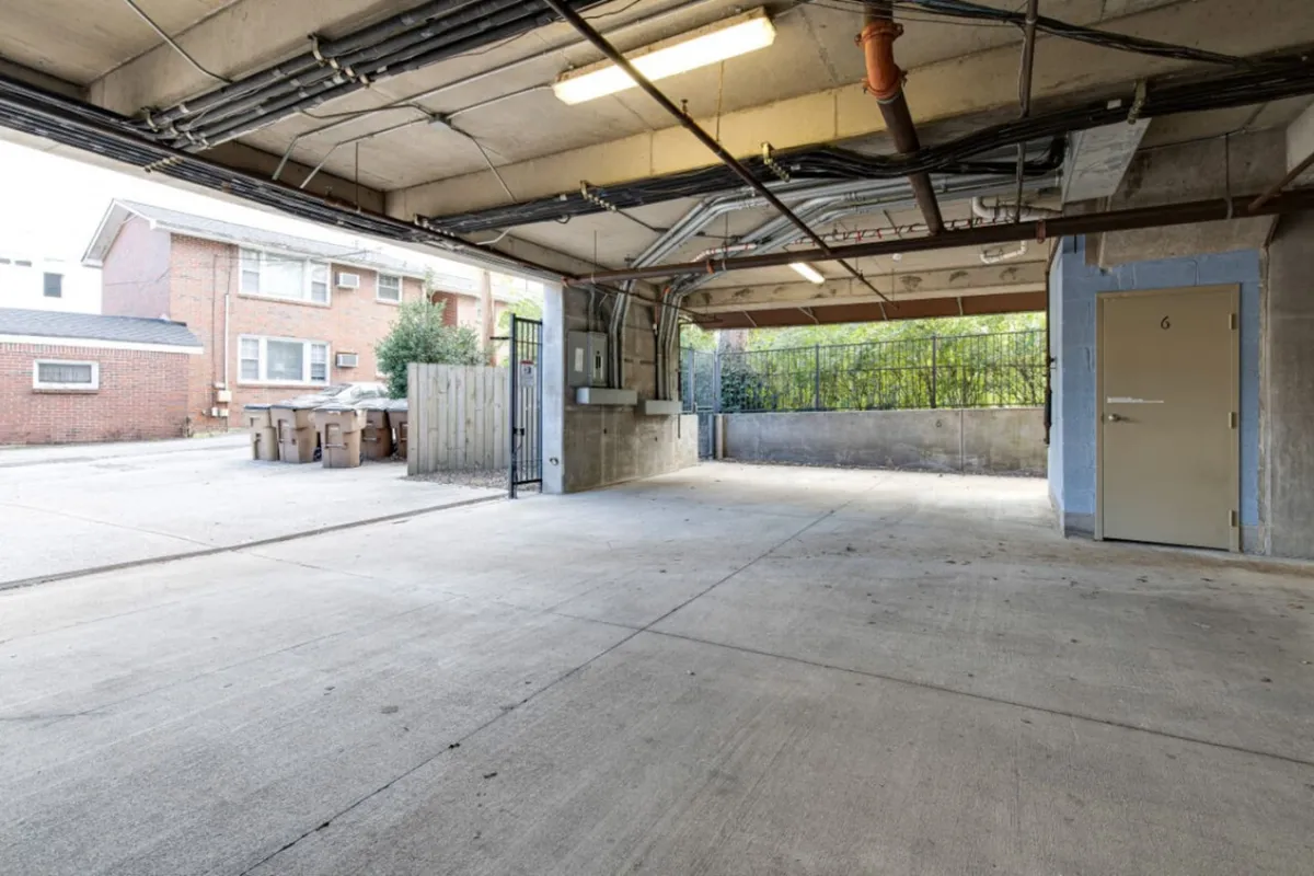 A practical garage environment featuring a concrete floor, open parking space, exposed pipes, and a visible doorway to the building. The setup offers straightforward access for residents as well as possibilities for community interaction in the shared garage space.