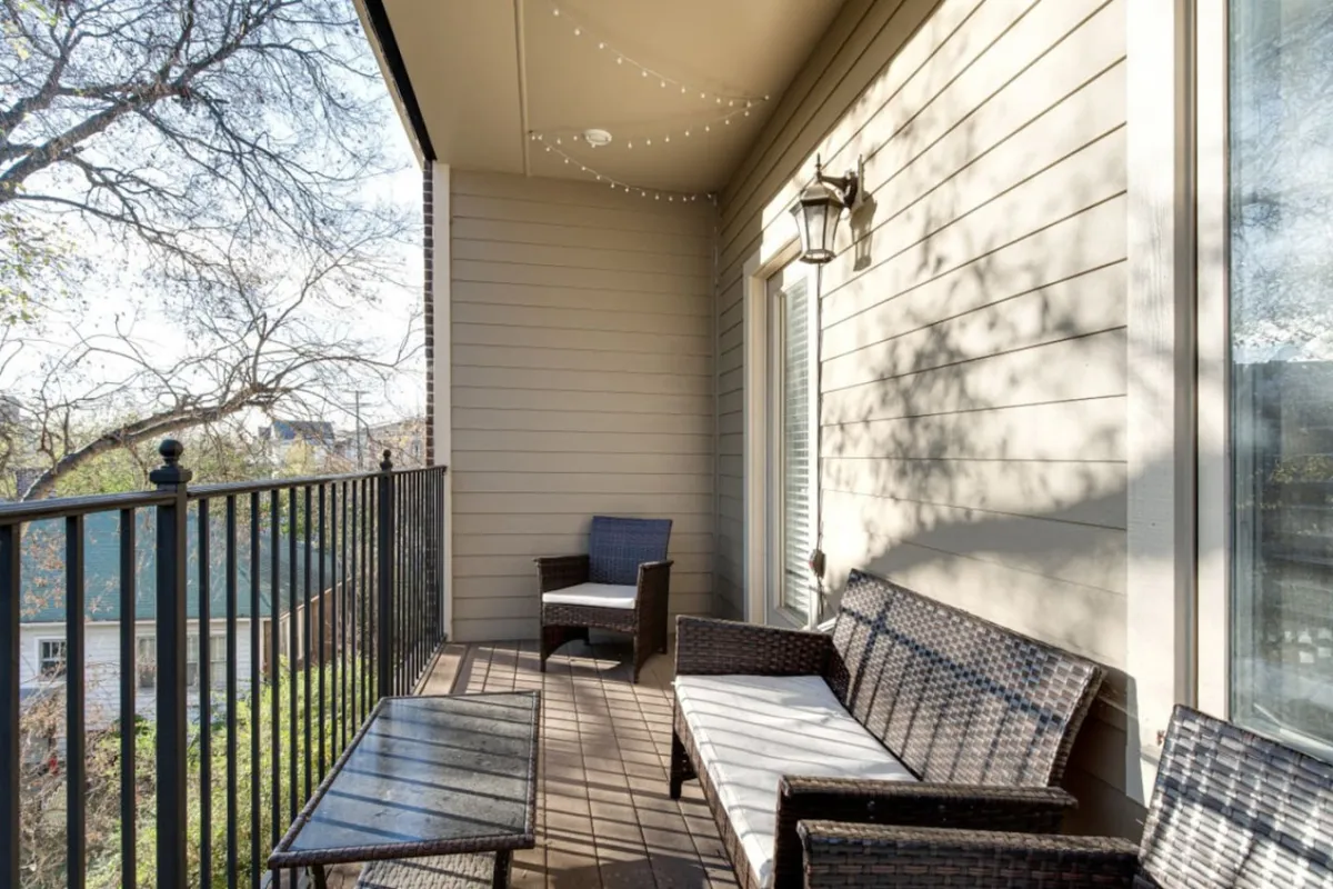 An inviting exterior balcony space featuring comfortable wicker seating, a sturdy balcony railing, and twinkling string lights overhead. The atmosphere is serene and welcoming, perfect for relaxing after a day spent exploring local attractions. The elevated vantage point gives a glimpse of the surrounding environment, suggesting easy access to the neighborhood's parks and eateries.
