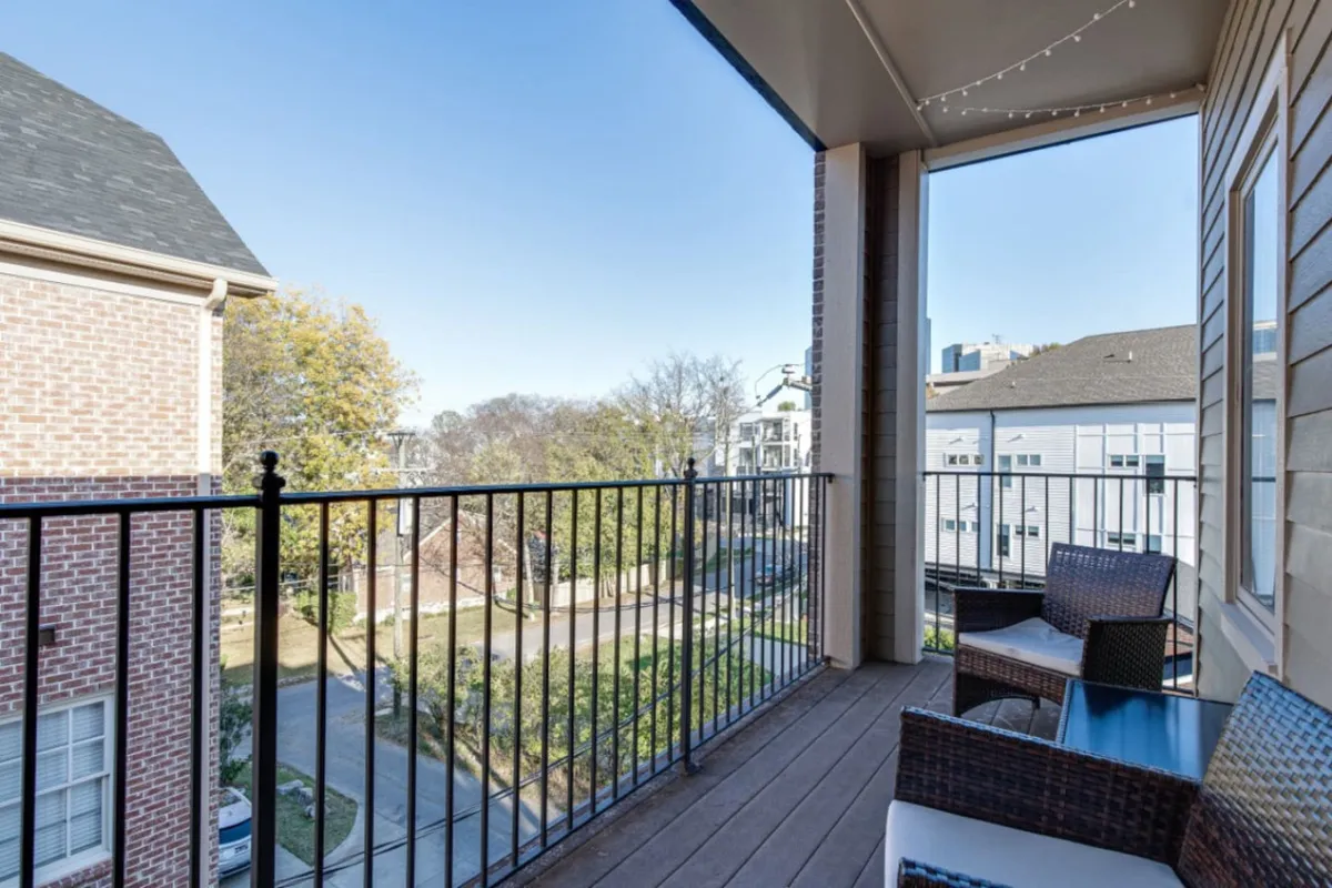 An inviting balcony with comfortable seating overlooks lush trees, capturing the tranquil transition from the private condo to the lively city beyond. The string lights and outdoor setup hint at small gatherings and moments of relaxation before or after a night out.
